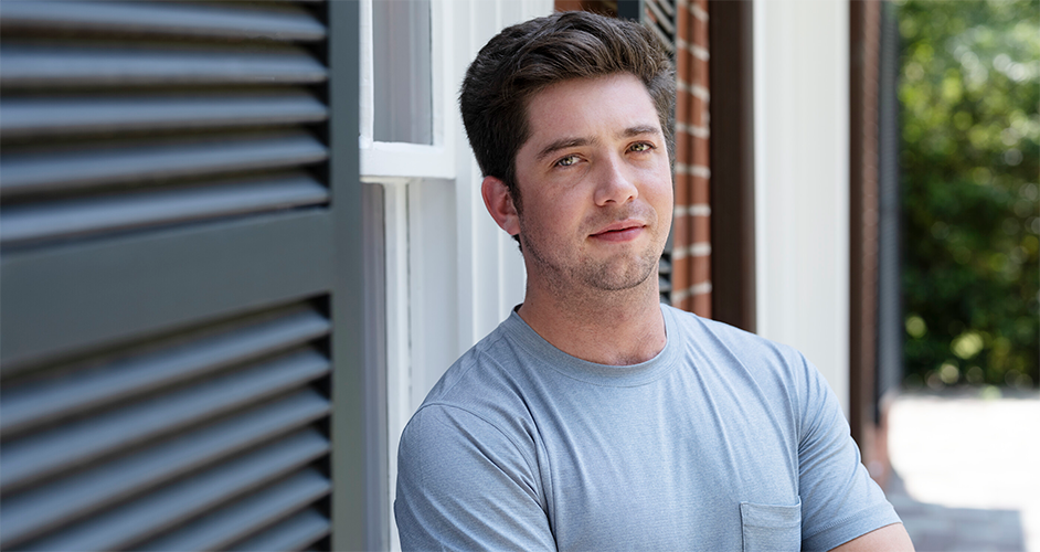 A young man named Freeman stands outdoors in a casual gray t-shirt, leaning against a wall with arms crossed. The setting includes a building with white and brown siding, adjacent to lush greenery.