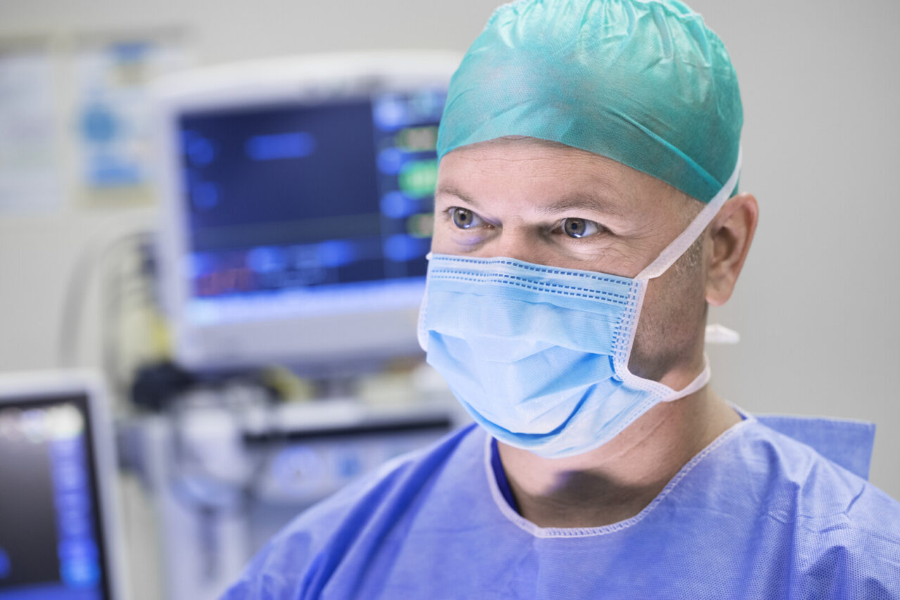 A medical professional in surgical attire, including a blue gown, mask, and a green surgical cap, is in a hospital setting. In the background, there are medical monitors and equipment, indicating an operating room environment.