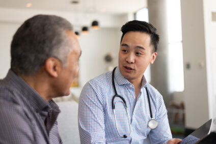 A doctor of Asian descent and an elderly patient are indoors at the man's home. The doctor is talking to his patient about his health while showing him his tablet.